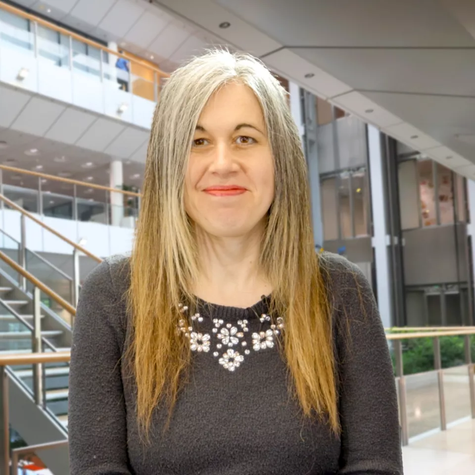 Female with long grey/blonde hair standing in an office atrium smiling 