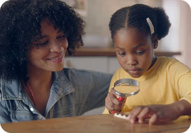 Woman smiling while child examines a Kinder Chocolate with a magnifying glass at a table.