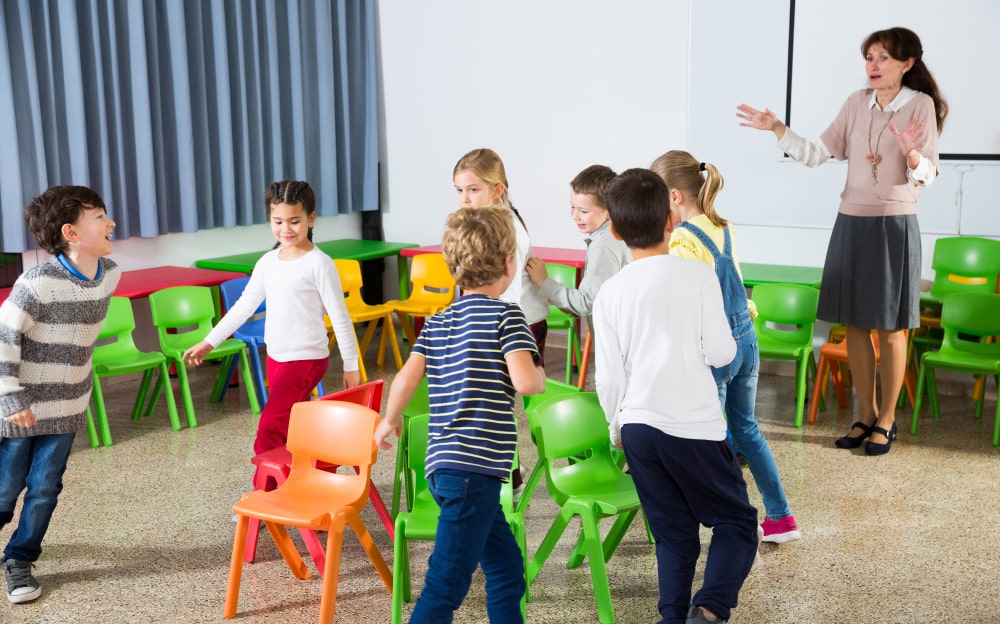 Group of children moving around chairs as teacher gives instructions.