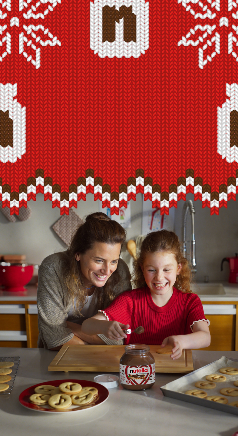 A young girl is using a spoon to scoop Nutella® out of a jar to decorate Nutella® biscuits, while her mother watches over her shoulder.